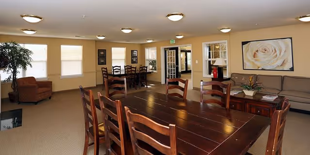 A spacious common area with a large wooden dining table surrounded by chairs in the foreground. In the background, there is a smaller table with chairs near windows with blinds. To the right, there is a beige couch with a wooden coffee table and a large framed picture of a white rose on the wall. The room has beige walls, carpeted floor, and ceiling lights.
