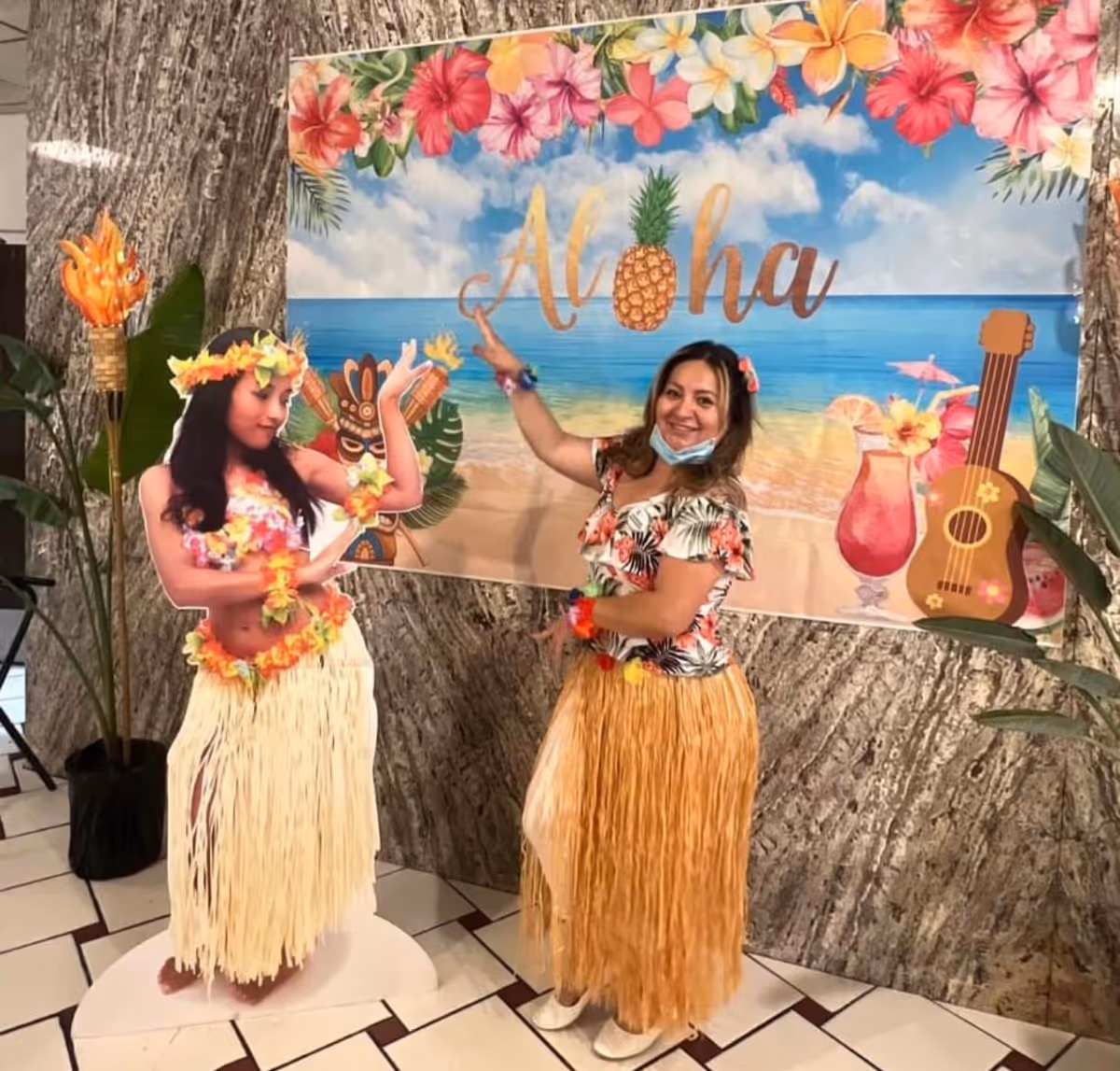 A woman dressed in a floral top and a grass skirt poses next to a life-sized cutout of a hula dancer wearing a flower lei and grass skirt. Behind them is a colorful backdrop with tropical flowers, a beach scene, and the word 'Aloha' with a pineapple replacing the letter 'o'. There are also tropical decorations like a tiki torch and large green leaves.