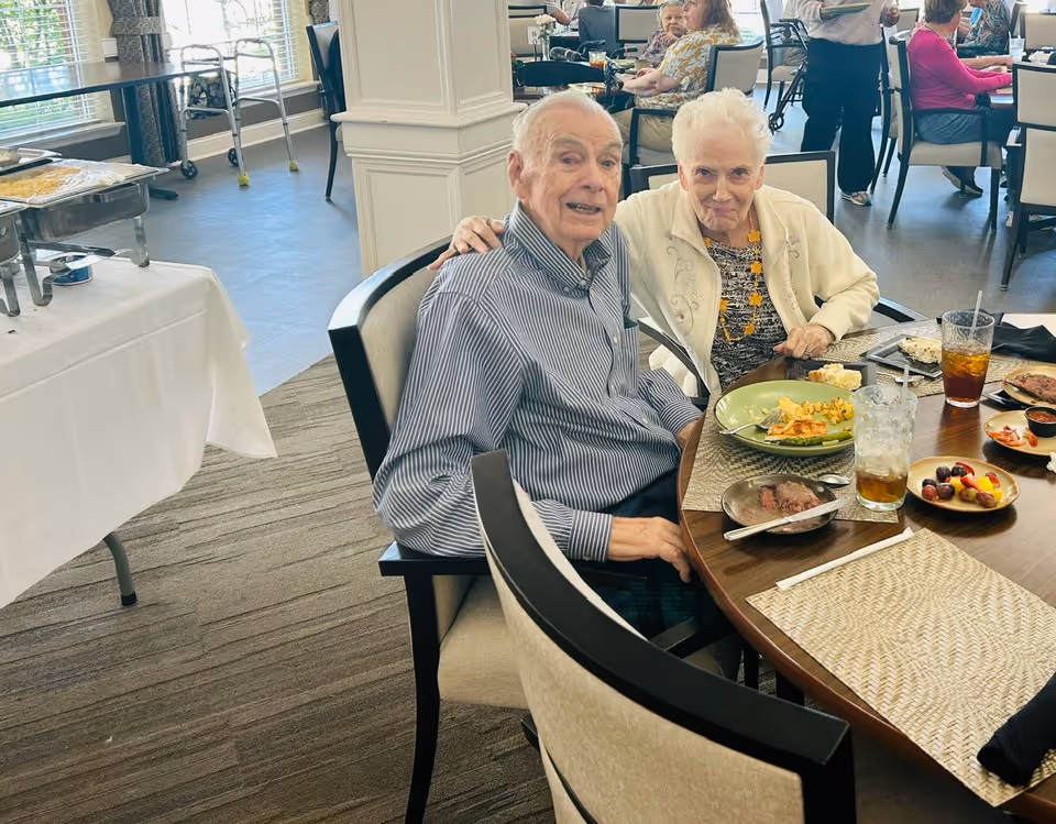 An elderly man and woman sitting together at a round dining table in a senior living facility dining room. They have plates of food and drinks in front of them. Other residents are visible in the background also seated at tables and eating. The room has large windows with blinds and a buffet table with food on the left side.