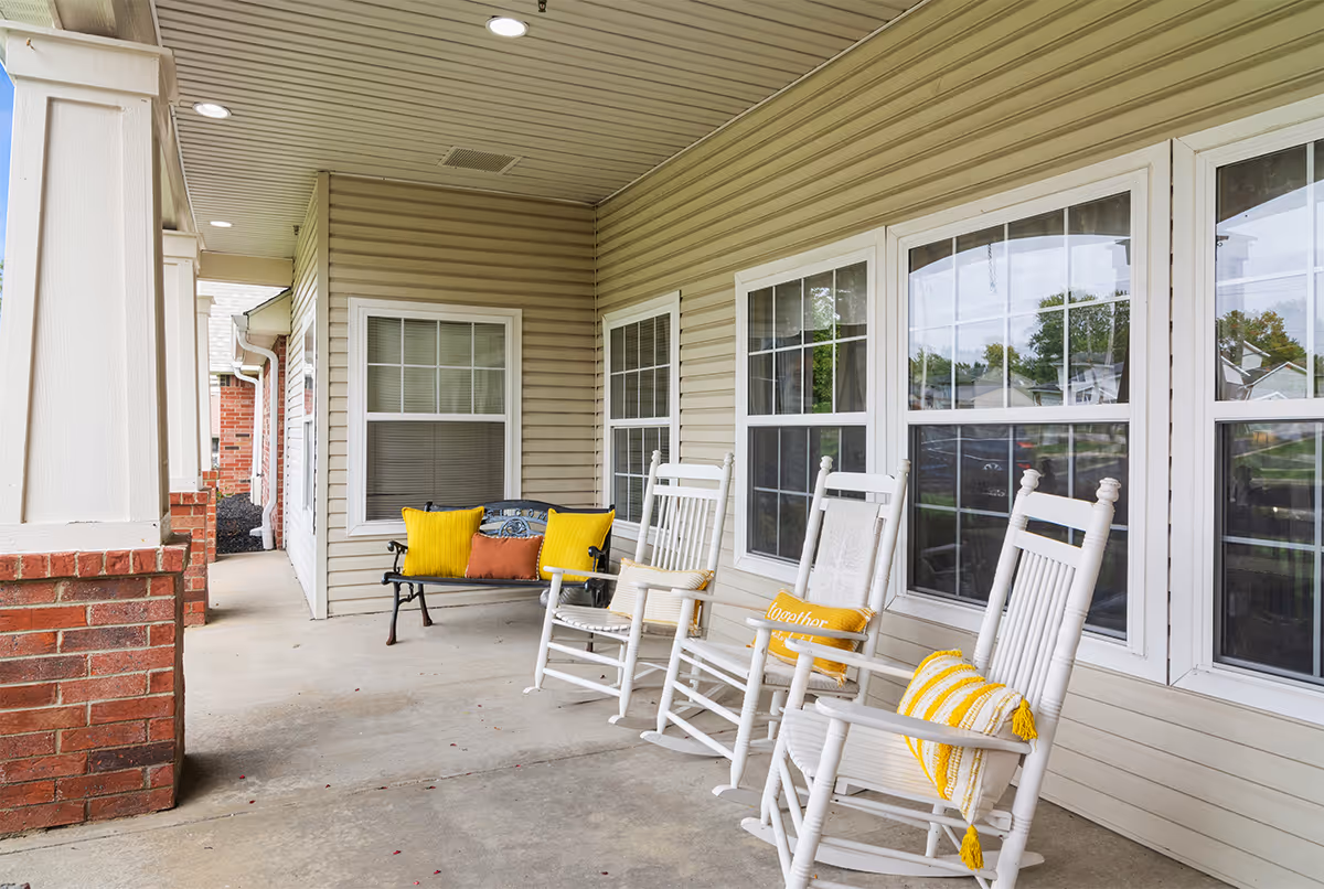 Covered outdoor porch area with three white rocking chairs each with yellow and white cushions, and a black metal bench with yellow and orange cushions. The porch has beige siding, white framed windows, and a brick column.