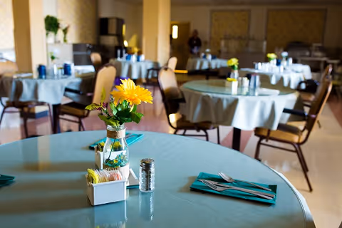 A bright dining room with round tables covered in light blue tablecloths, each set with teal napkins, silverware, and small flower vases with yellow flowers. Chairs are arranged around the tables and the room has warm lighting.