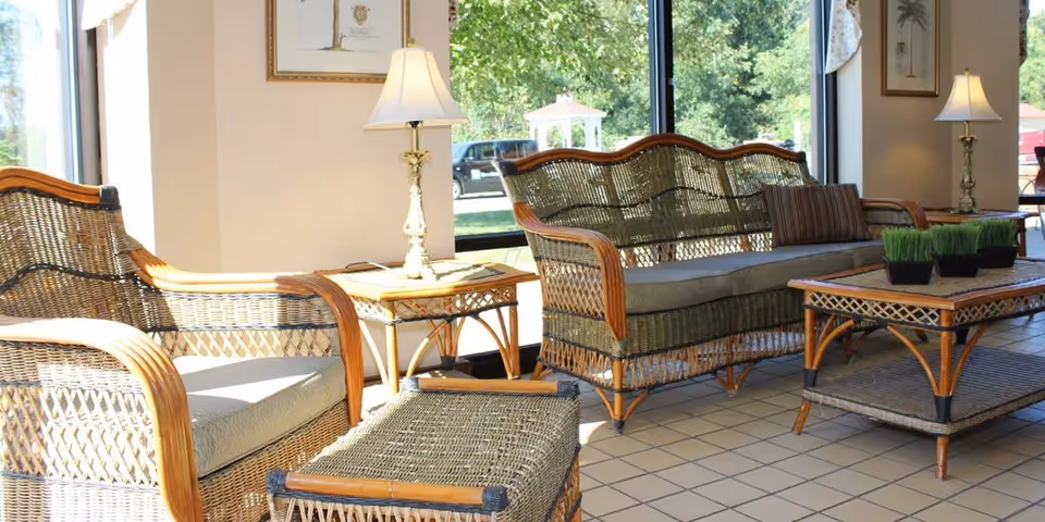 A bright sitting area with wicker furniture including a cushioned armchair, a cushioned loveseat, a coffee table with two small potted plants, and a side table with a lamp. Large windows in the background show greenery and a gazebo outside.