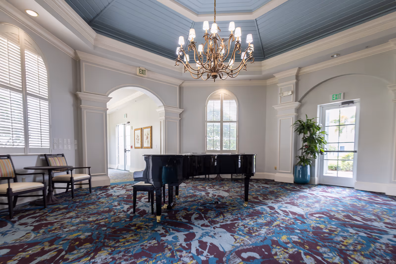 Bright octagonal lounge with a grand piano under a chandelier, patterned carpet, arched windows and seating.