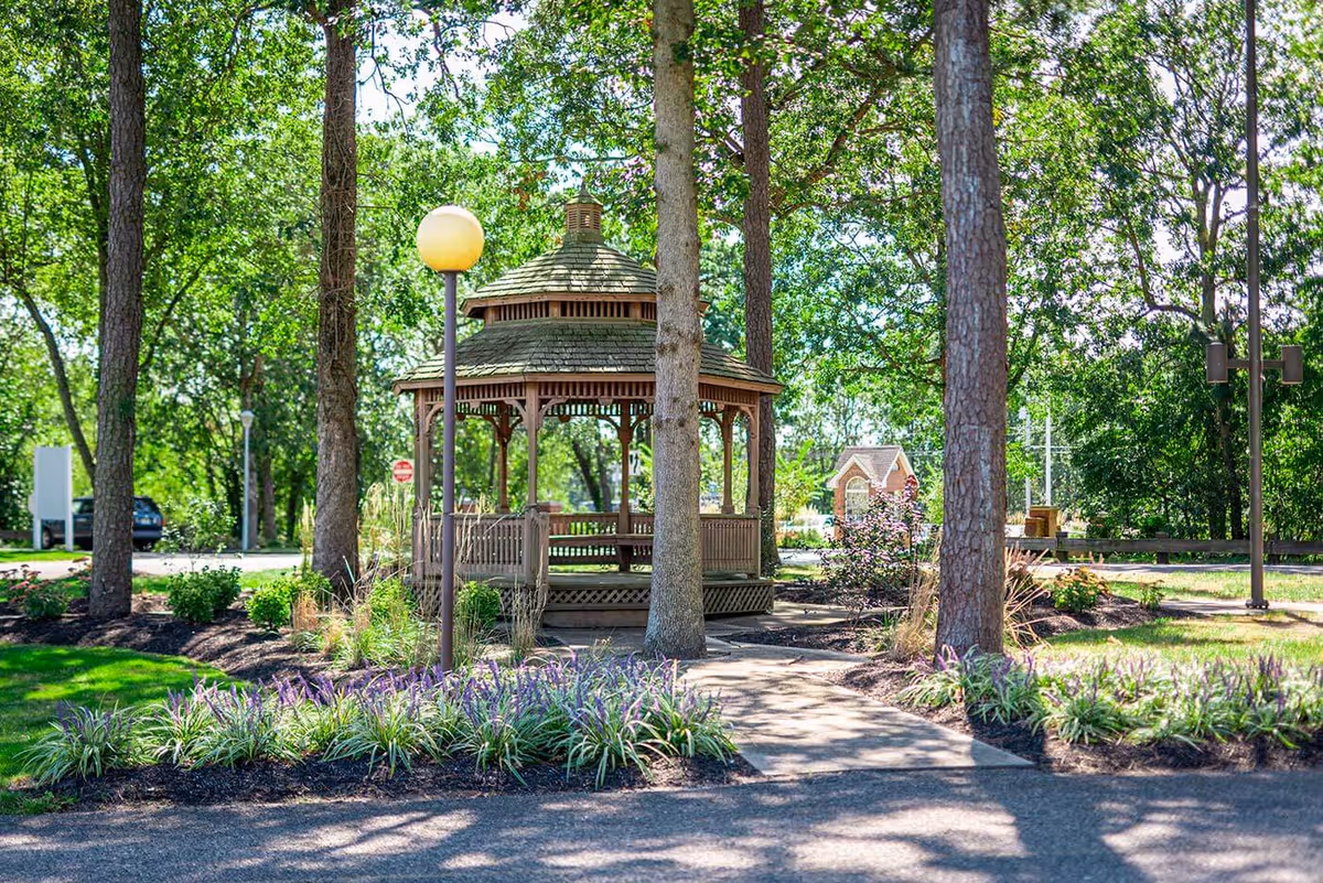 A wooden gazebo sits among trees and landscaped flower beds on a sunny outdoor path.