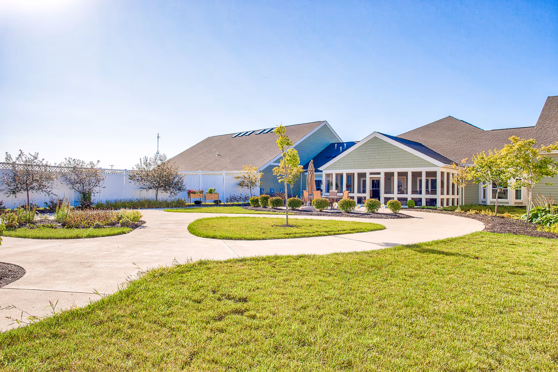 A bright outdoor courtyard area at Landings of Sidney featuring a curved concrete walkway surrounded by green grass, small trees, and landscaped garden beds. The building has light green siding with a screened porch area and several chairs visible. The sky is clear and blue.