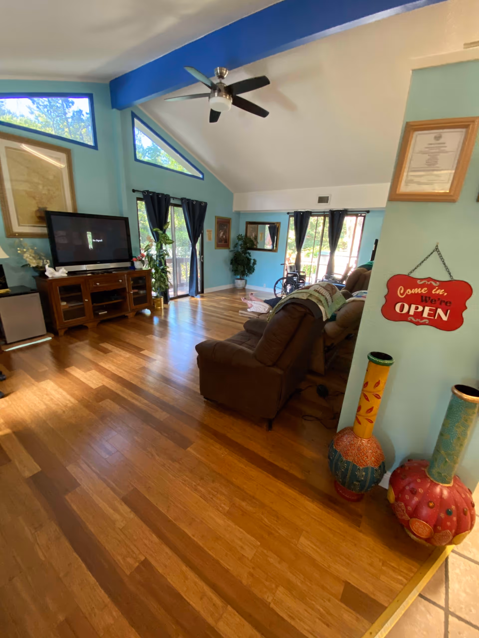 A bright living room with wooden floors, light blue walls, and a vaulted ceiling with a ceiling fan. There is a large flat-screen TV on a wooden stand, several windows with dark curtains letting in natural light, a brown recliner chair, and decorative vases near the entrance. A red sign on the wall reads 'Come in, We're OPEN'.