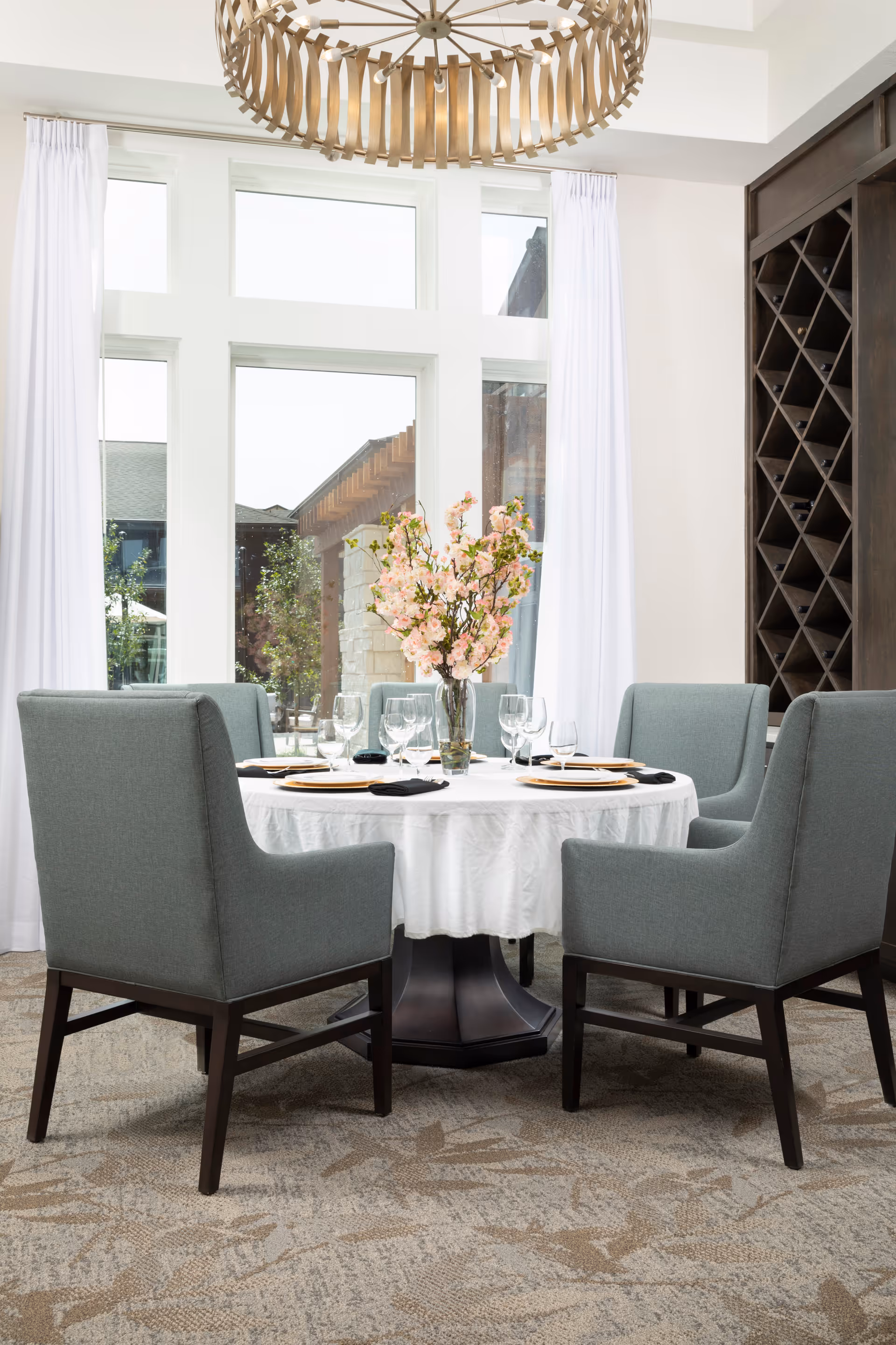 A round dining table covered with a white tablecloth set with plates, wine glasses, and black napkins, surrounded by four gray upholstered chairs. A vase with pink flowers is placed in the center of the table. Behind the table are large windows with white curtains, and to the right is a dark wooden wine rack. A modern chandelier hangs from the ceiling above the table.