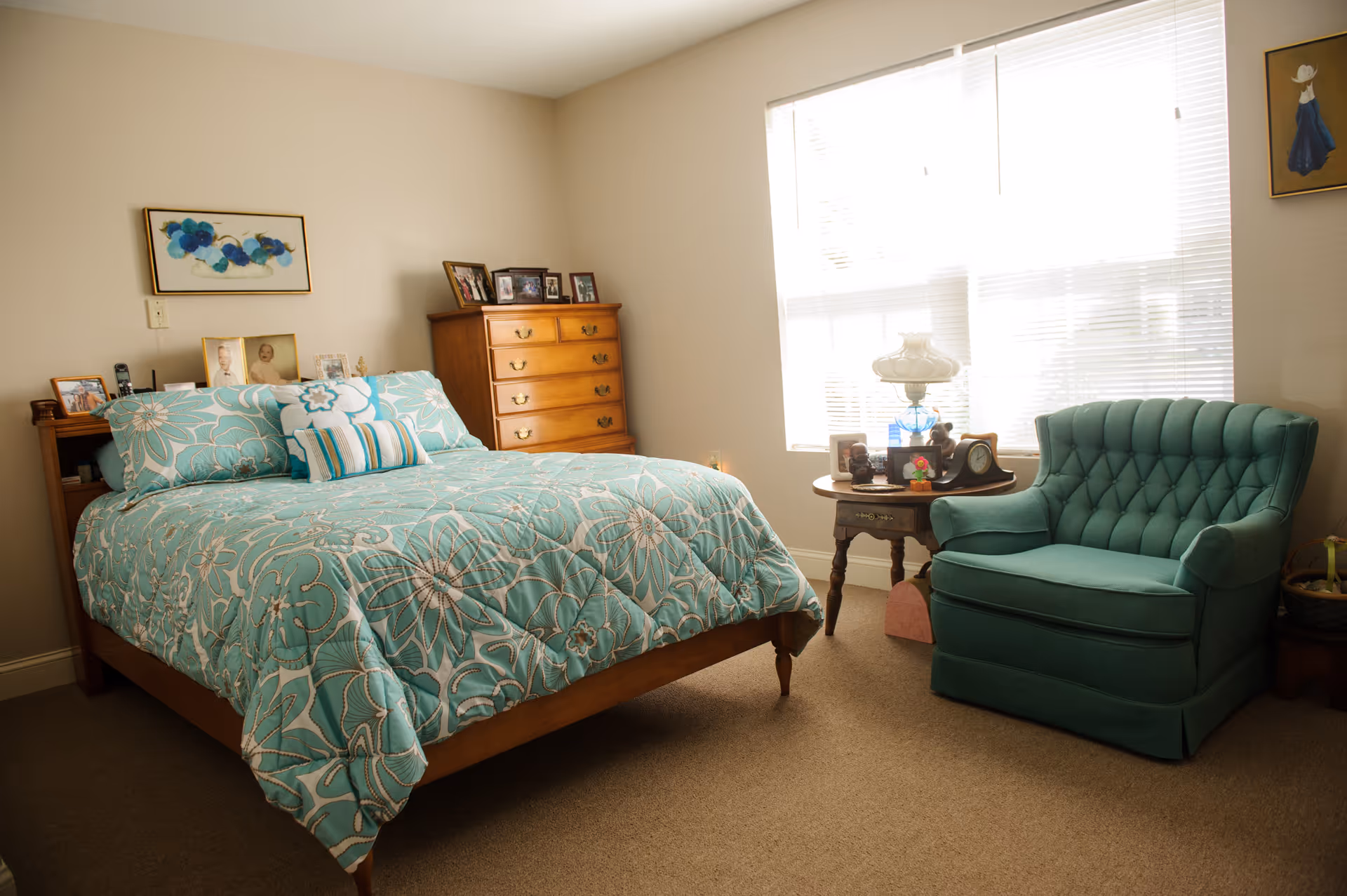 A cozy bedroom with a wooden bed covered in a teal floral comforter and matching pillows. Next to the bed is a wooden dresser with framed photos on top. A round wooden side table holds a decorative lamp, clock, and other small items. A teal upholstered armchair is positioned near a large window with blinds, allowing natural light to fill the room. The walls are decorated with framed artwork.
