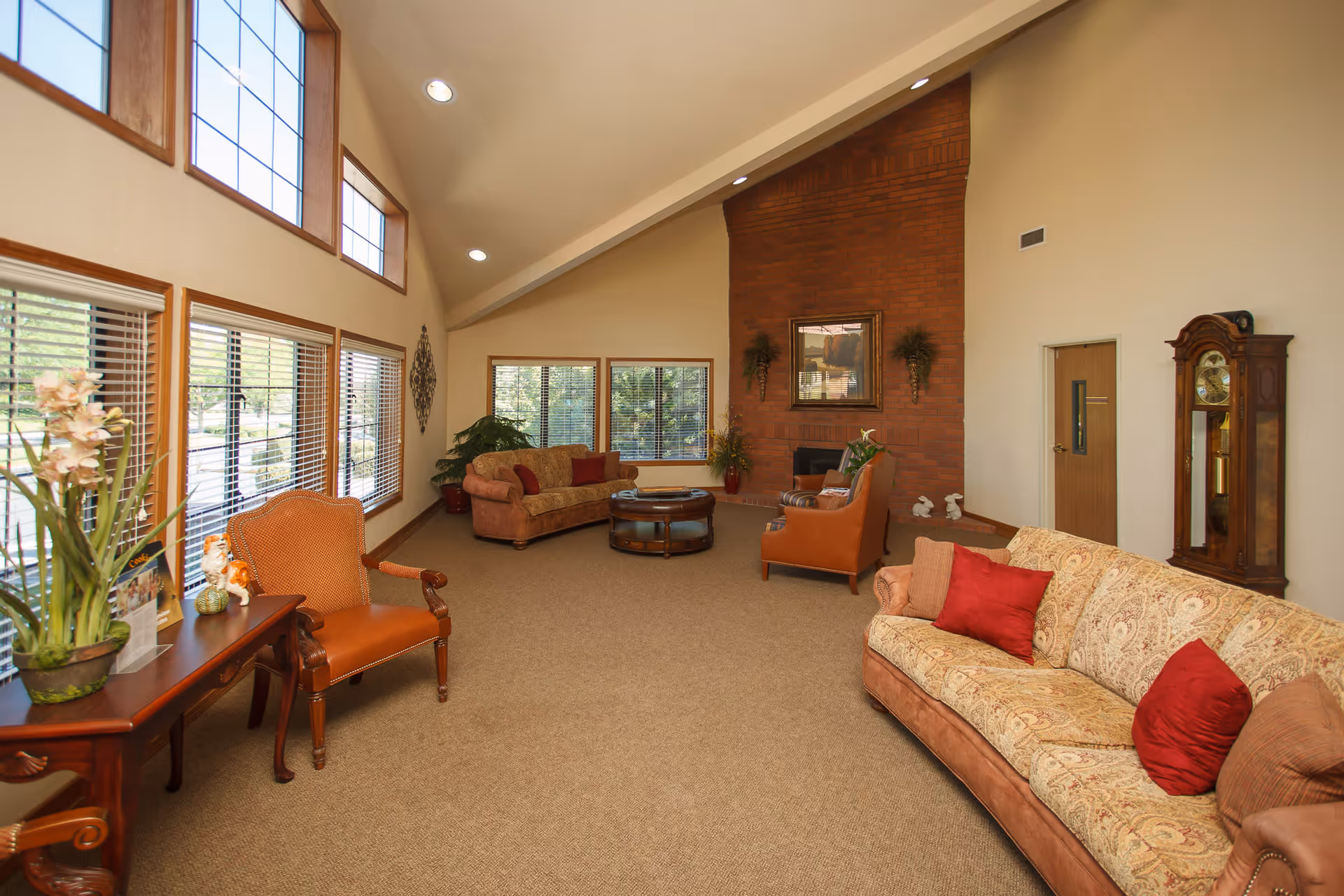 Spacious lounge with high ceilings, large windows, sofas and chairs arranged around a central coffee table and a brick fireplace.