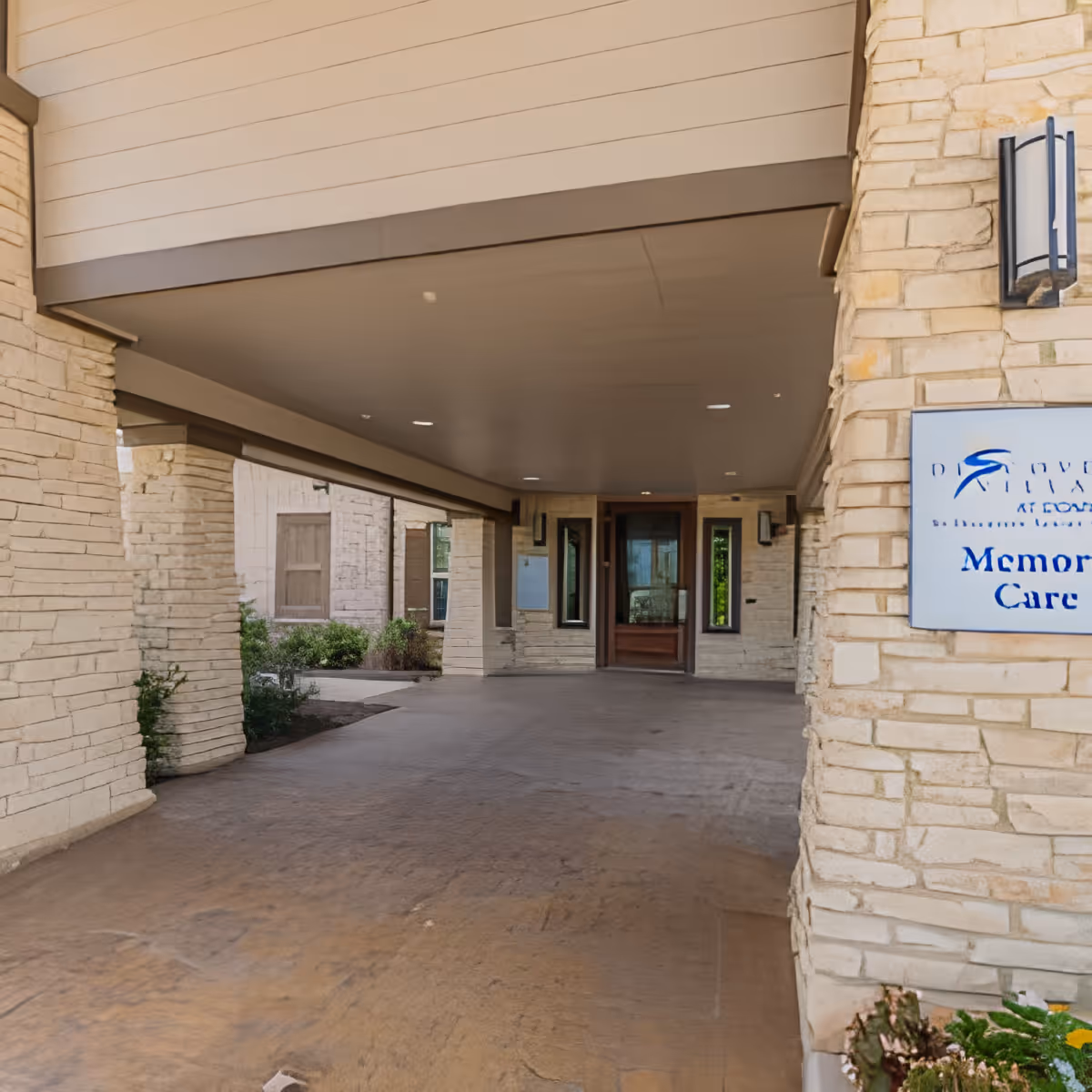 Covered entrance area of a building with stone walls and a sign indicating Memory Care at Discovery Village At Dominion. The entrance has a wide driveway and a wooden door at the end.