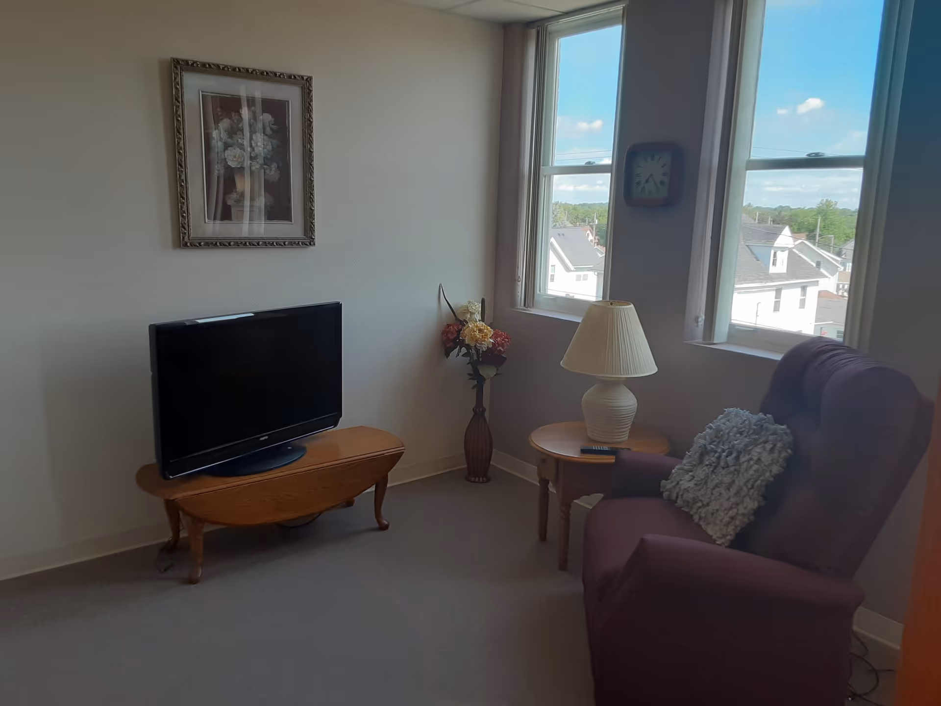 Small sitting area with a TV on a wooden table, an upholstered armchair with a throw pillow, a side table and lamp, a vase of flowers, and two windows showing rooftops.