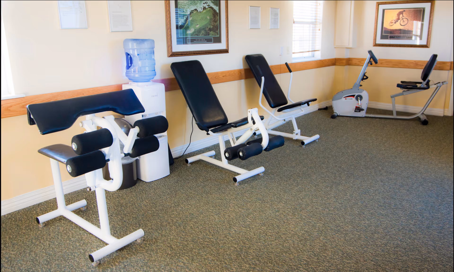 A small exercise room with three workout machines lined up against a beige wall with wooden trim. There is a water cooler between the first and second machines. Two framed pictures hang on the walls, and a window with blinds lets in natural light.