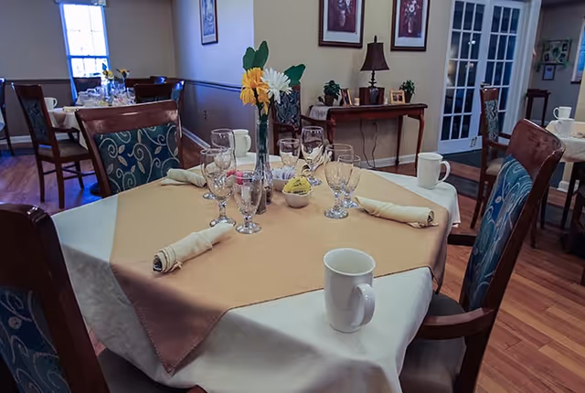A dining room table set with a beige tablecloth and a brown overlay, featuring neatly rolled napkins, multiple drinking glasses, a white coffee mug, and a small vase with yellow and white flowers. The room has wooden chairs with patterned cushions, wooden flooring, framed pictures on the wall, and a side table with a lamp and decorative items in the background.