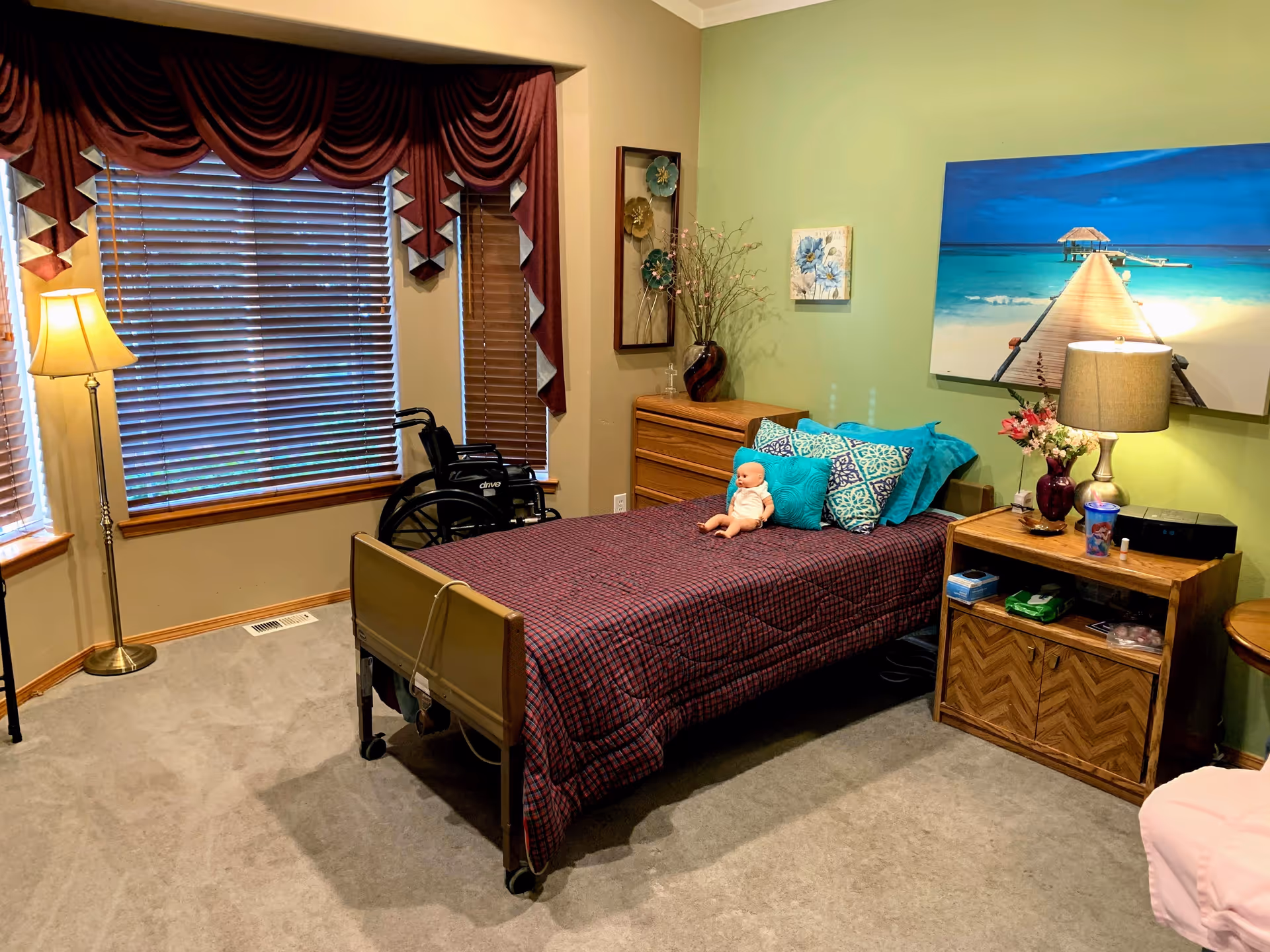 Cozy single-occupancy bedroom with a hospital-style bed, decorative pillows and a doll, a wheelchair by the window, dresser and nightstand with lamp and beach artwork on the wall.