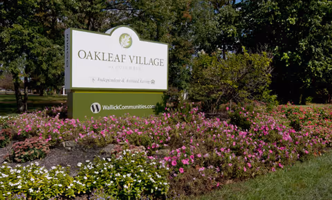 A large sign for Oakleaf Village of Columbus surrounded by a landscaped garden with pink and white flowers and green bushes, set against a backdrop of trees and grass under a clear sky.