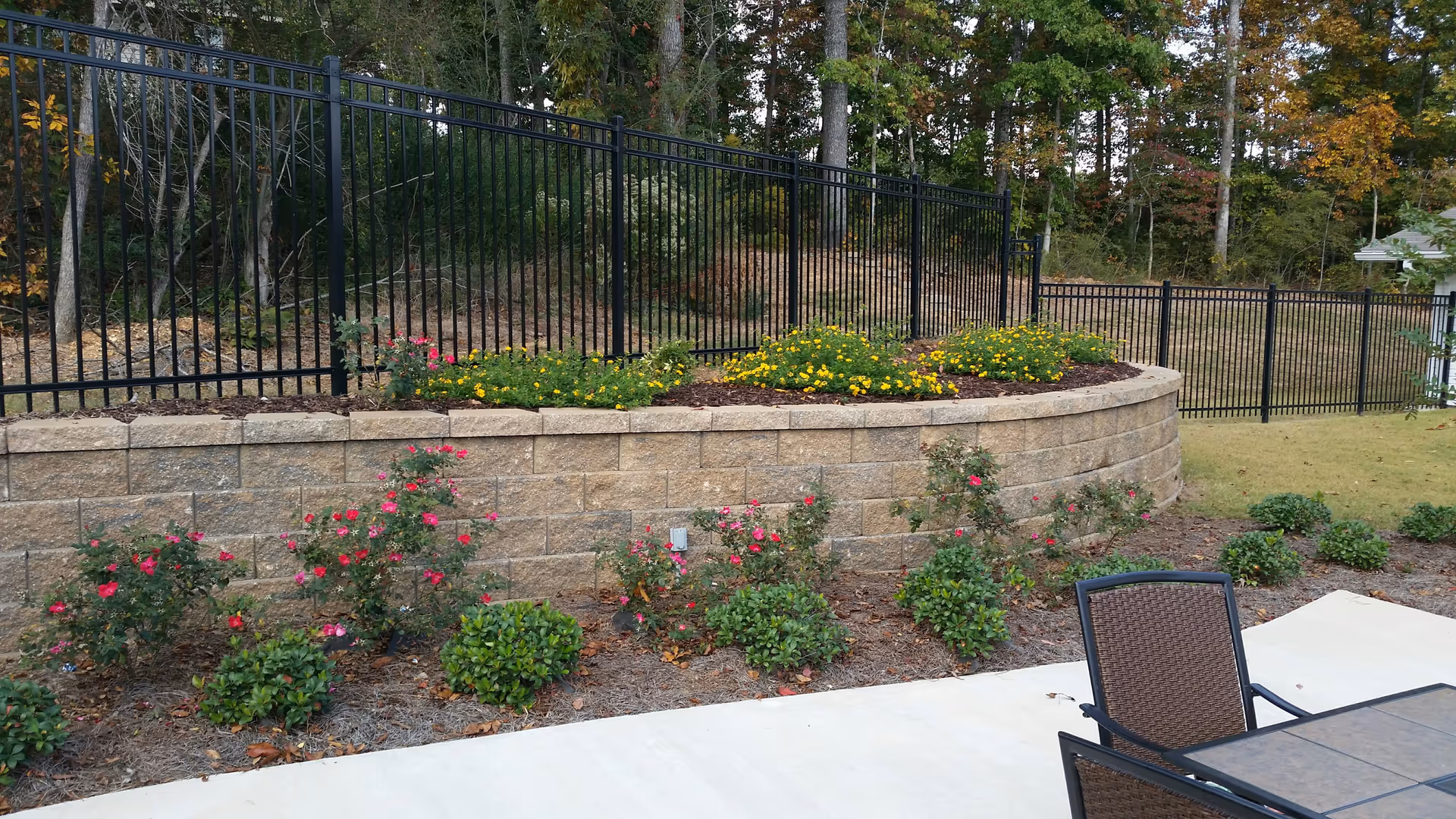 Outdoor patio area with a metal table and chairs on a concrete surface, bordered by a landscaped garden with bushes and flowering plants. Behind the garden is a retaining wall made of stone blocks and a black metal fence, with trees and greenery in the background.