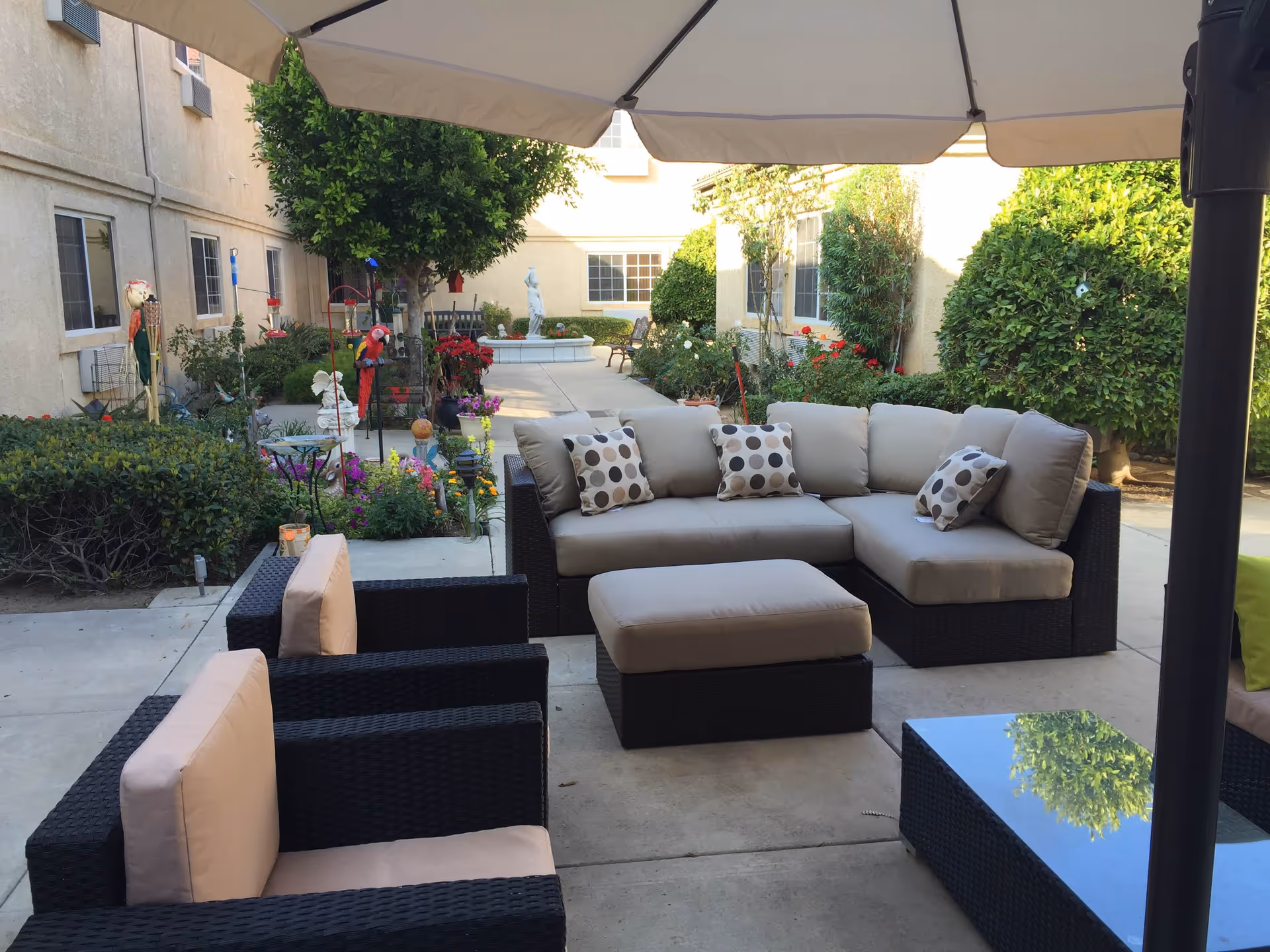 Outdoor patio area with cushioned wicker seating including a sectional sofa, two armchairs, and an ottoman under a large umbrella. The patio is surrounded by greenery, flowers, and bushes with a building in the background.