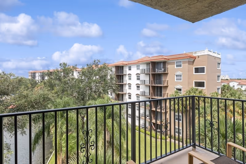View from a balcony with a railing overlooking palm trees, a pond and a neighboring multi-story apartment building under a blue sky.