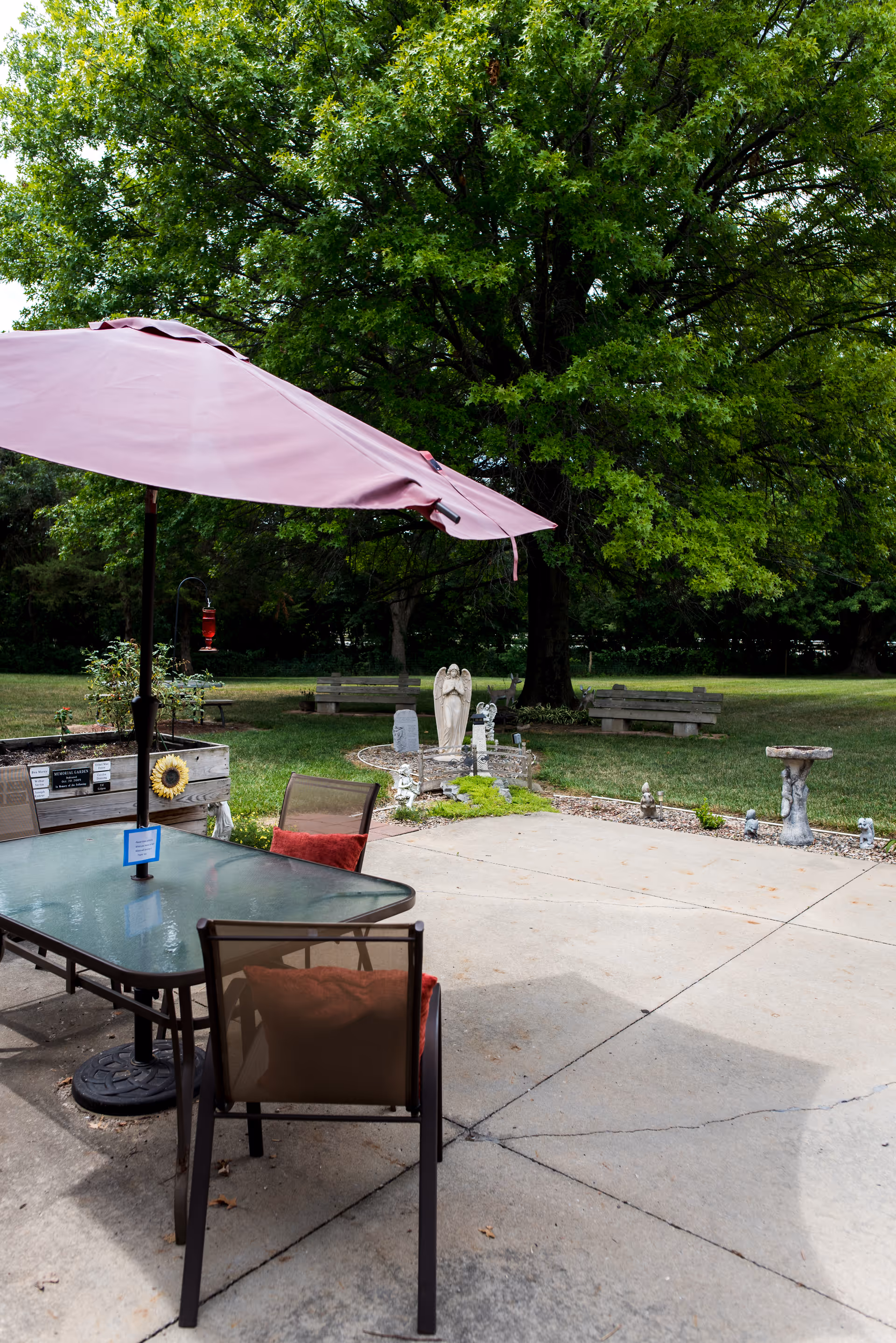 Outdoor patio area with a glass table and chairs under a large pink umbrella. The patio is surrounded by green grass, trees, benches, and garden decorations including a statue of an angel and a birdbath.