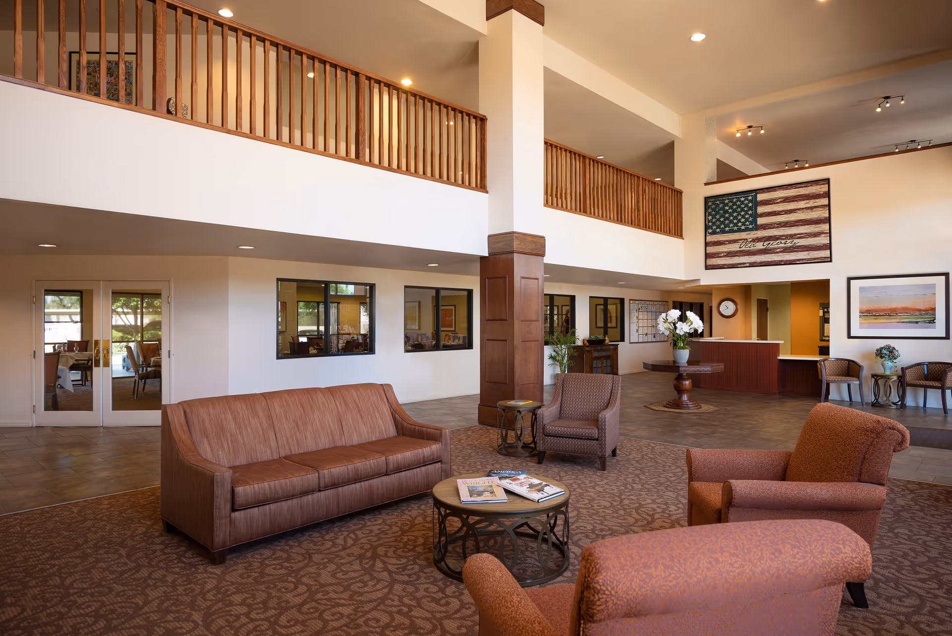 Spacious assisted living facility common area with brown upholstered sofa and armchairs arranged around a round coffee table with magazines. The room features a high ceiling with wooden railings on the upper level, a reception desk in the background, and decorative wall art including a large American flag and a framed landscape painting.