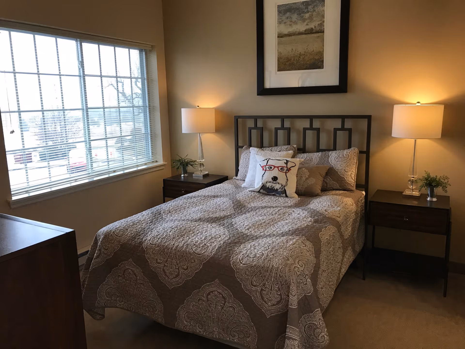 A cozy bedroom with a double bed covered in a patterned beige and white quilt. The bed has several pillows, including one with a dog wearing red glasses. There are two bedside tables, each with a lamp and a small plant. A large window with blinds is on the left wall, and a framed picture hangs above the bed.