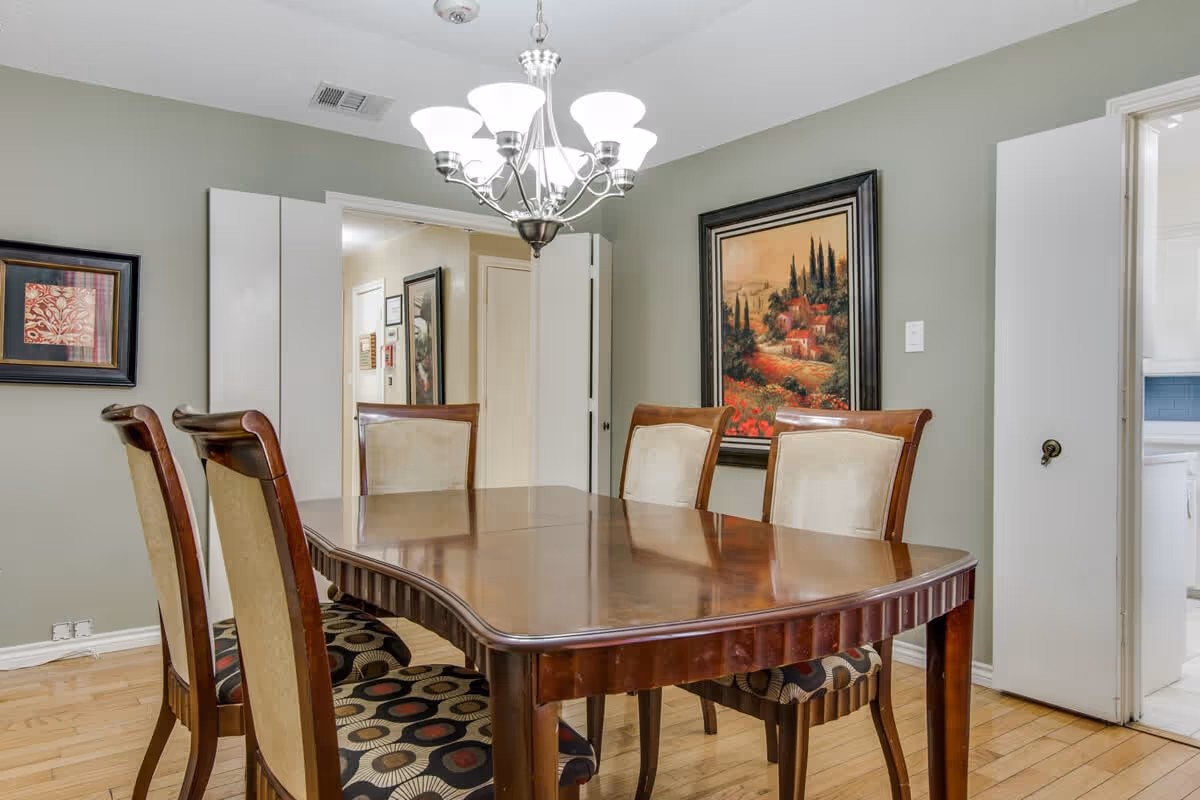A dining room with a polished wooden table and six chairs, four with beige upholstery and two with patterned cushions. The room has light green walls adorned with two framed paintings. A chandelier with multiple lights hangs above the table. There is a doorway leading to a hallway and another doorway opening into a kitchen area with white cabinets.