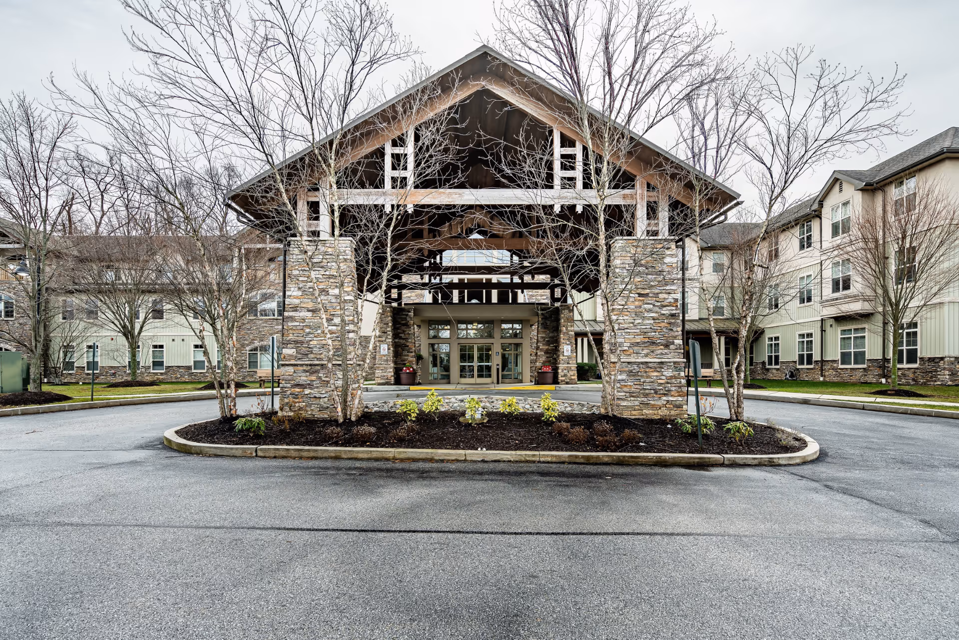 Front entrance and covered porte-cochère of the Spring Mill senior living building with stone pillars, a circular driveway, and leafless trees.