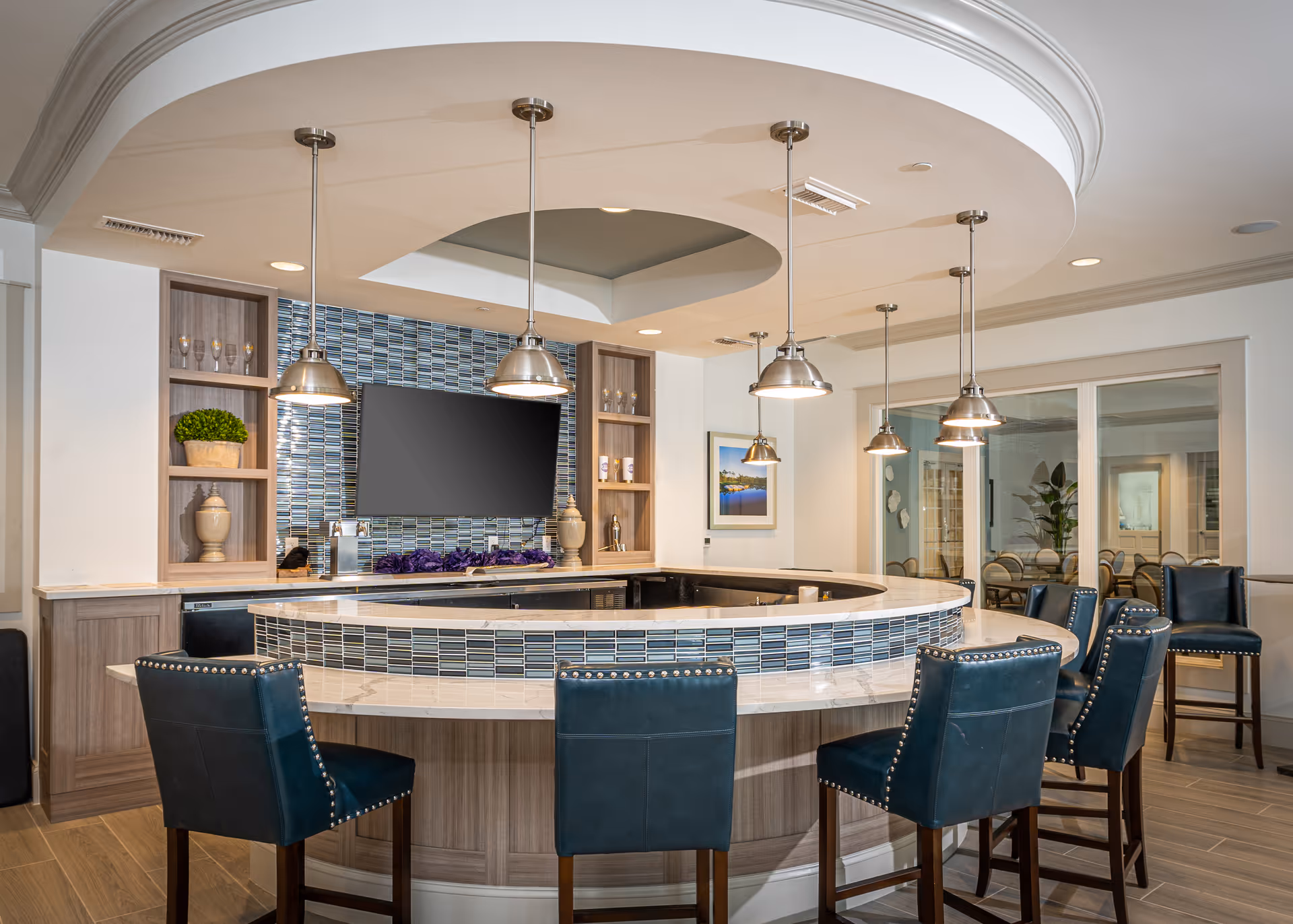 A modern curved bar area with a marble countertop and blue tiled front. Six dark blue leather bar stools with nailhead trim are arranged around the bar. Above the bar hang six metallic pendant lights. Behind the bar is a wall with blue mosaic tiles, open shelving with decorative items, and a mounted flat-screen TV. The room has wood flooring and a large window showing another room with chairs and a plant.