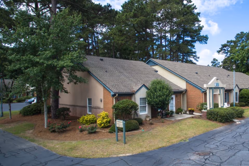 Exterior view of a single-story residential building surrounded by trees and shrubs with a paved driveway in front under a partly cloudy sky.