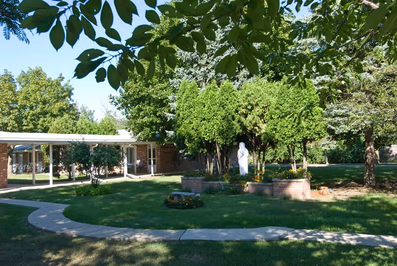 Outdoor garden area at Clinton Villa with a curved concrete walkway, green grass, various trees, and a white statue surrounded by brick planters with flowers.