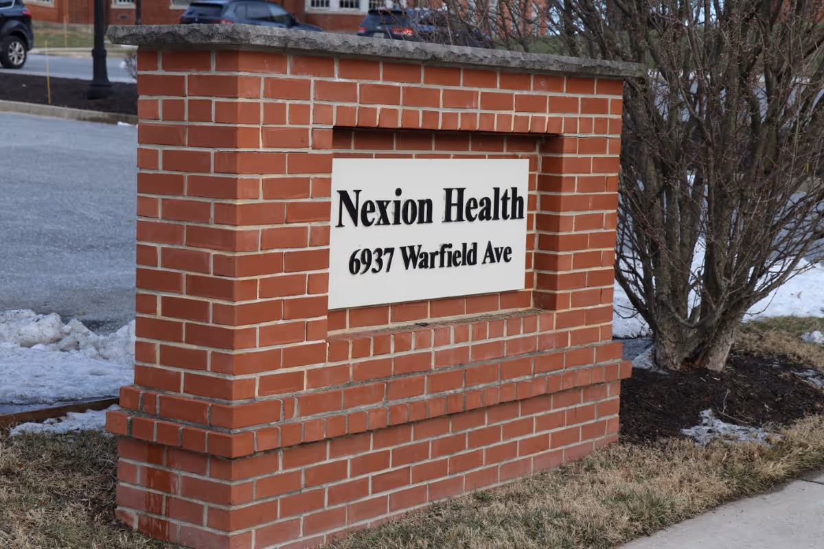 A brick signpost with a white plaque displaying the text 'Nexion Health 6937 Warfield Ave' located outdoors near a sidewalk and some leafless bushes.