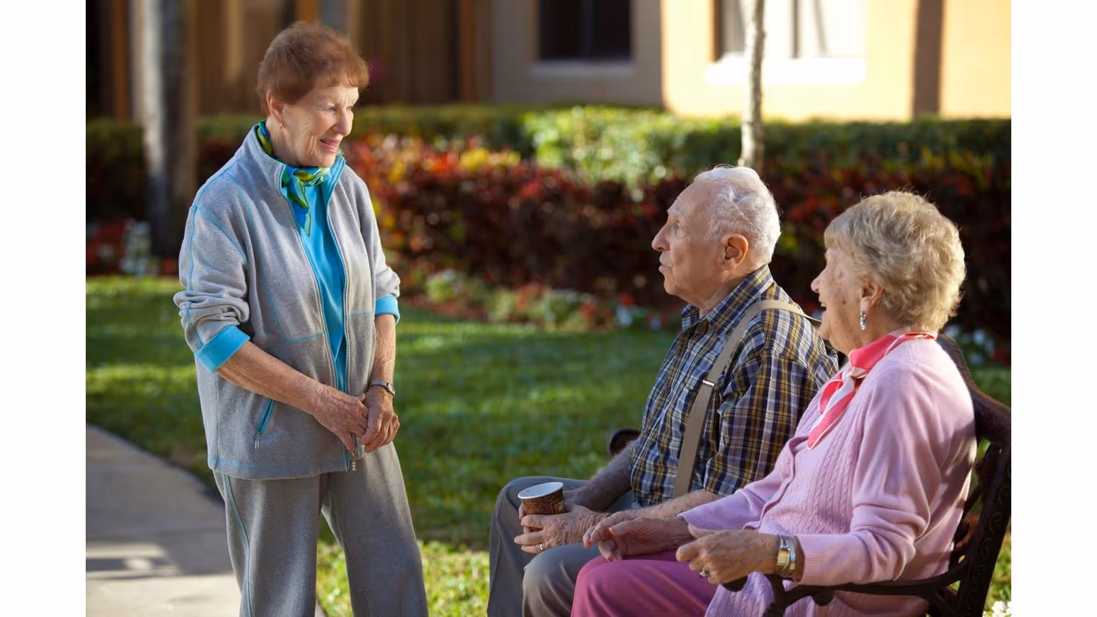 An elderly woman in a light gray tracksuit and blue scarf stands and smiles while talking to an elderly man and woman sitting on a bench outdoors in a garden area with green grass and shrubs in the background.