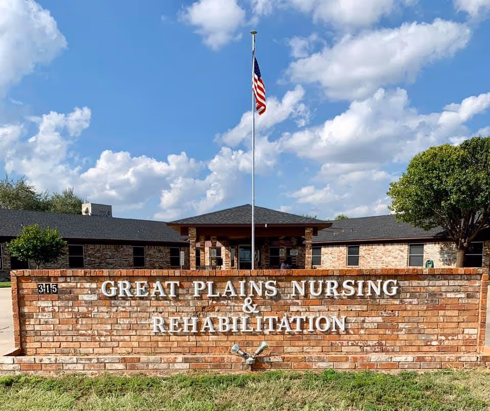 Exterior view of Great Plains Nursing and Rehabilitation facility showing a brick sign with the facility name in white letters, a flagpole with an American flag, and a building with a dark roof and brick walls under a partly cloudy blue sky.