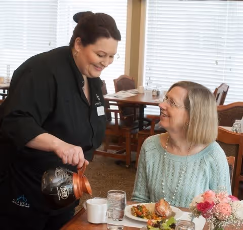A smiling staff member in a black uniform pours coffee into a cup for an elderly woman seated at a dining table with a plate of food and a glass of water. The setting is a dining room with wooden chairs and tables, and large windows with blinds in the background.
