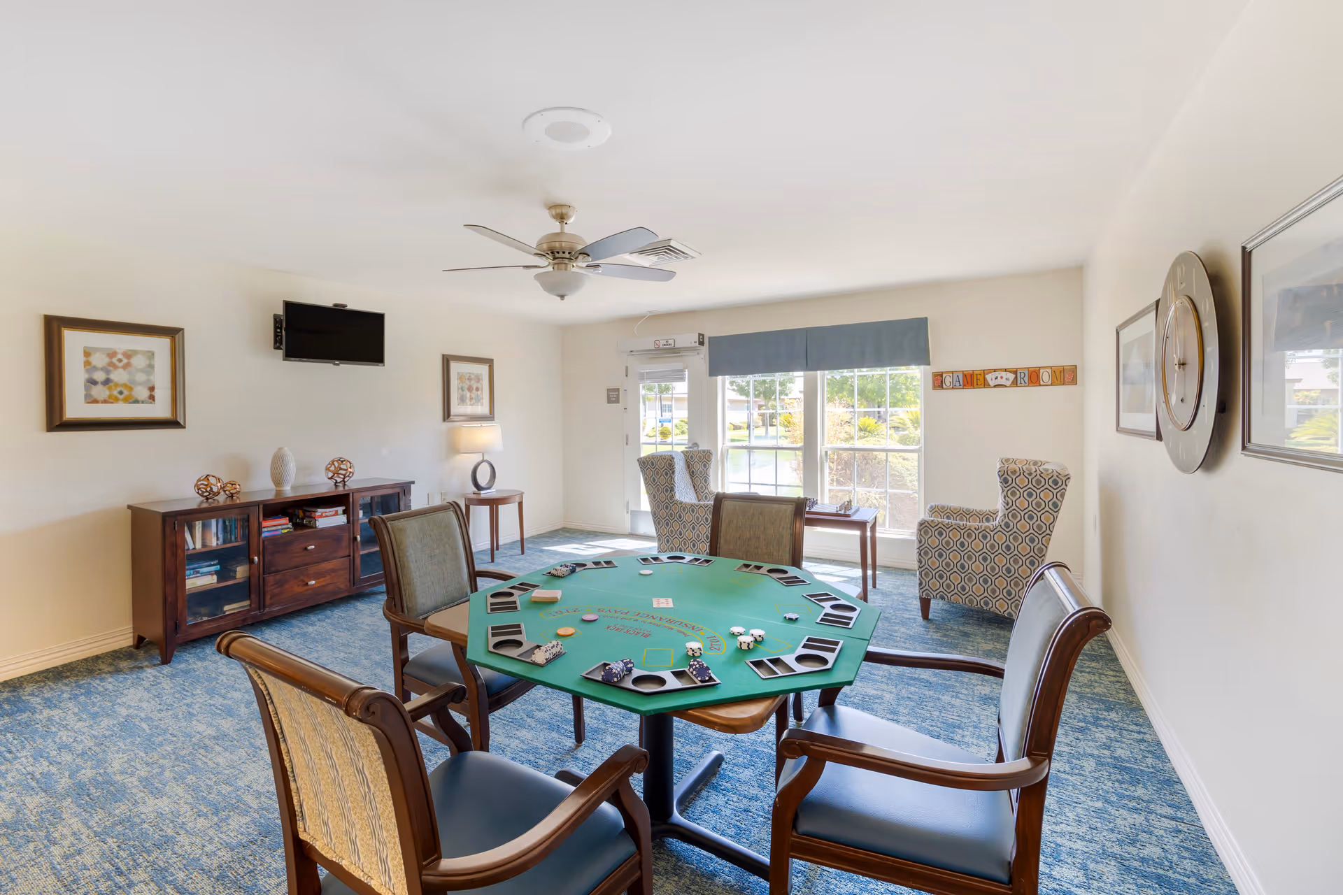 A bright game room with a green octagonal card table surrounded by four wooden chairs with cushions. The table has card holders and poker chips on it. The room features blue carpet, a ceiling fan, a wall-mounted TV, framed artwork, a wooden cabinet with books and decorative items, and two patterned armchairs near a large window with a blue valance. A sign on the wall reads 'GAME ROOM'.