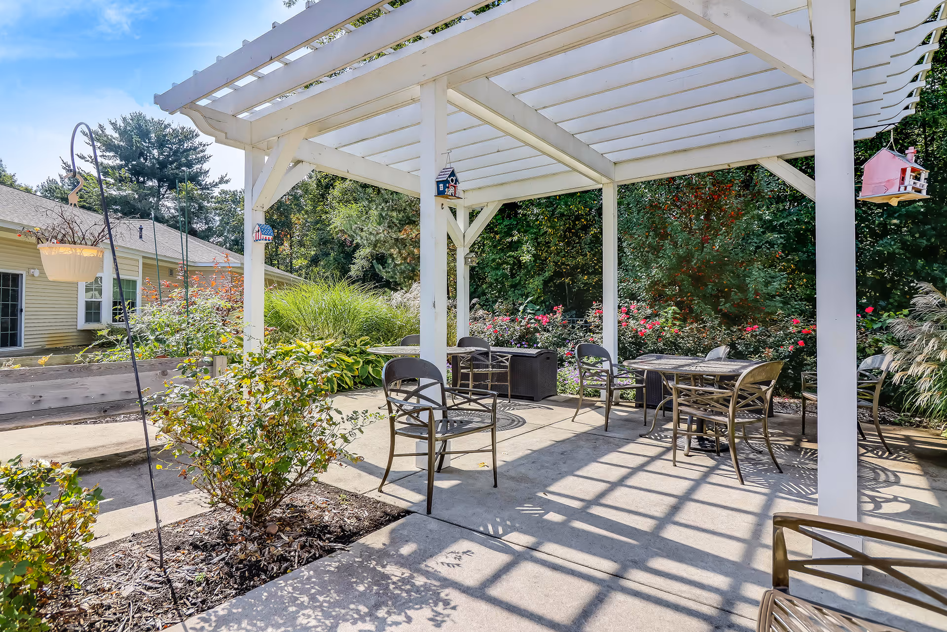 Outdoor patio area with a white pergola providing partial shade over several metal chairs and tables. Surrounding the patio are various plants and bushes, with a building visible in the background under a clear blue sky.