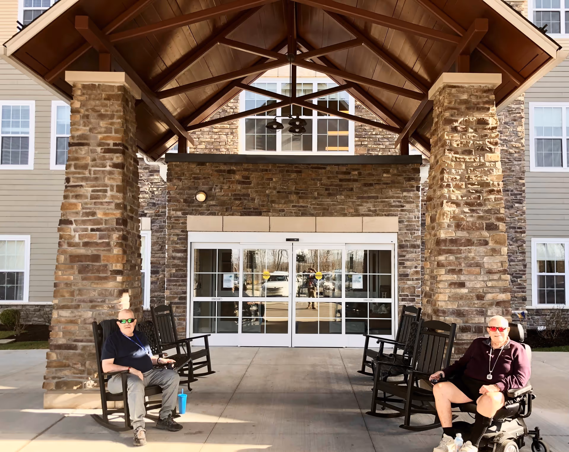Two elderly men sitting outside the entrance of a building with stone pillars and a wooden roof. One man is seated in a rocking chair and the other in a motorized wheelchair. The building has large glass double doors and multiple windows.