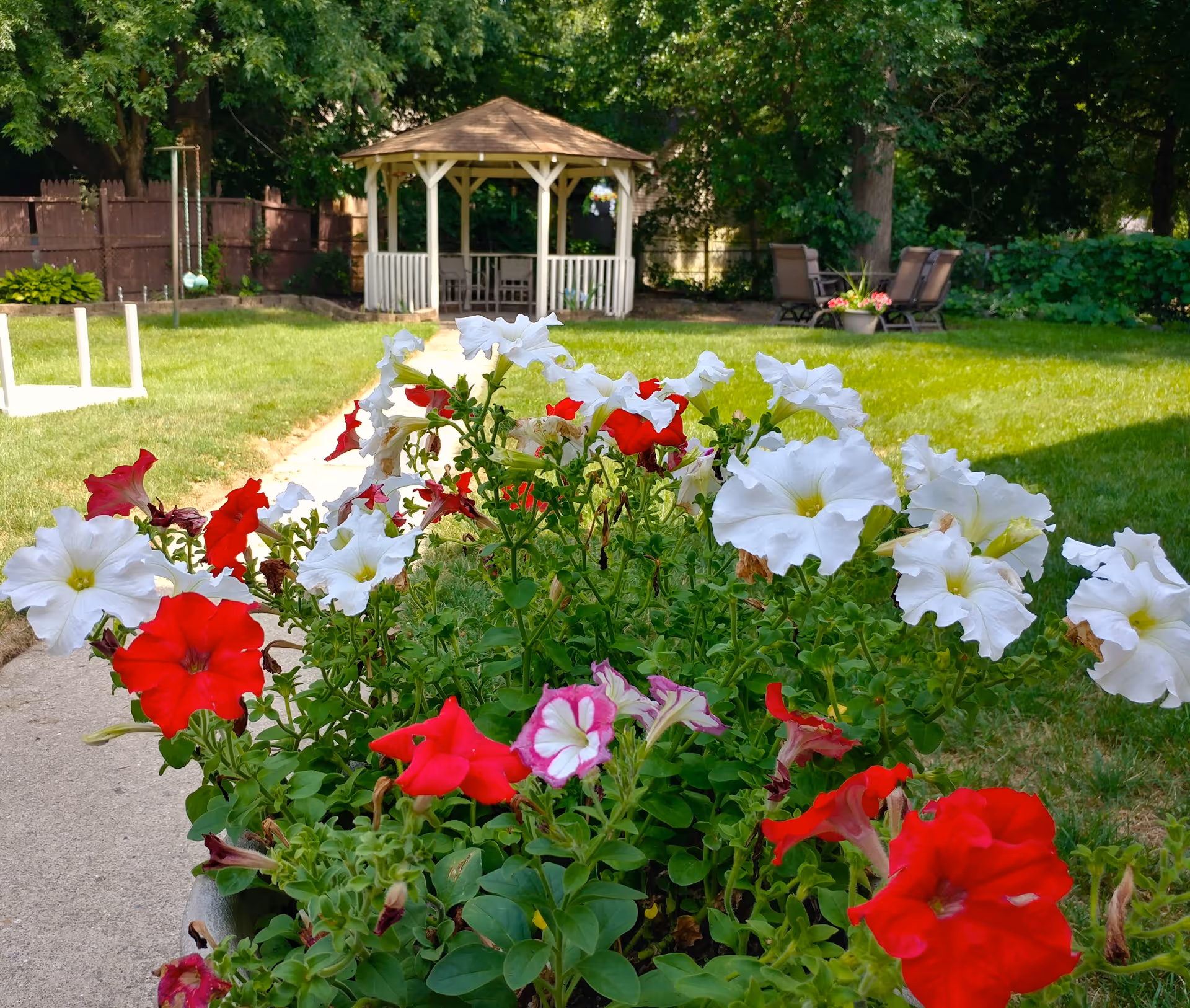 A garden area with vibrant red, white, and pink flowers in the foreground. In the background, there is a white gazebo surrounded by green grass, trees, and outdoor chairs. The scene is bright and sunny, suggesting a peaceful outdoor space.