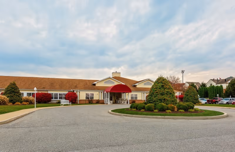 Front exterior view of a single-story senior living facility with a red canopy over the entrance, surrounded by landscaped bushes and a circular driveway under a cloudy sky.