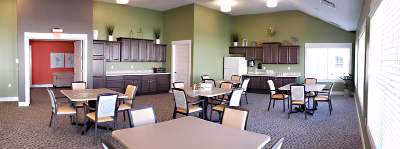 Bright multipurpose dining room with several tables and chairs, a kitchenette with cabinets and a refrigerator along green walls, and large windows.