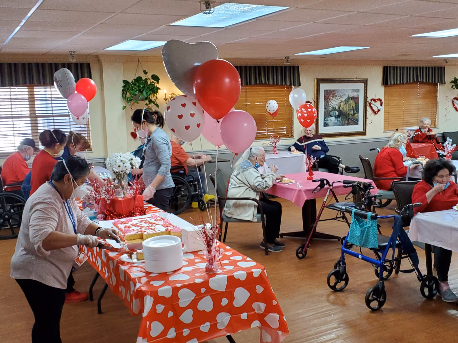 A group of elderly people and staff in a decorated common room at Mount Washington Care Center. The room has tables covered with red and pink heart-themed tablecloths and balloons, suggesting a Valentine's Day celebration. Some elderly residents are seated at tables eating, while staff members are serving cake. Walkers and wheelchairs are visible, and the room has warm lighting with framed artwork and plants on the walls.