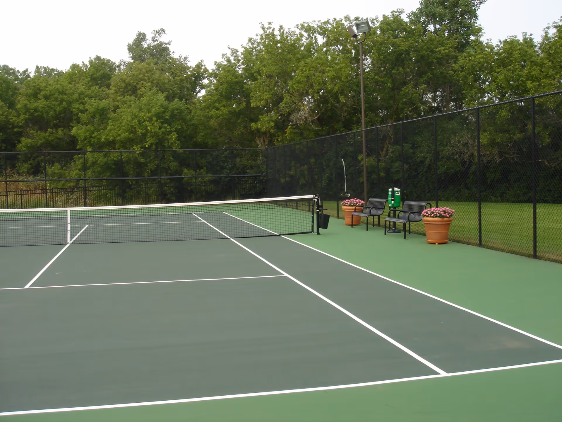 Outdoor tennis court with a net in the middle, surrounded by a black chain-link fence. There are two black benches, a green ball dispenser, and two large flower pots with pink flowers on the right side. Trees and greenery are visible in the background.