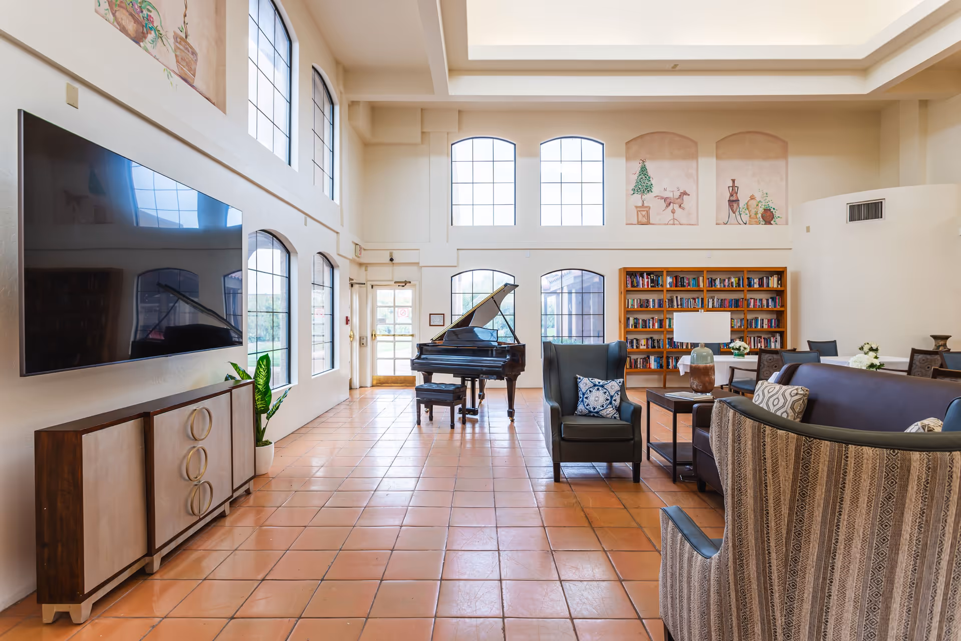 Spacious common room with a grand piano, seating area, bookshelves and large arched windows.