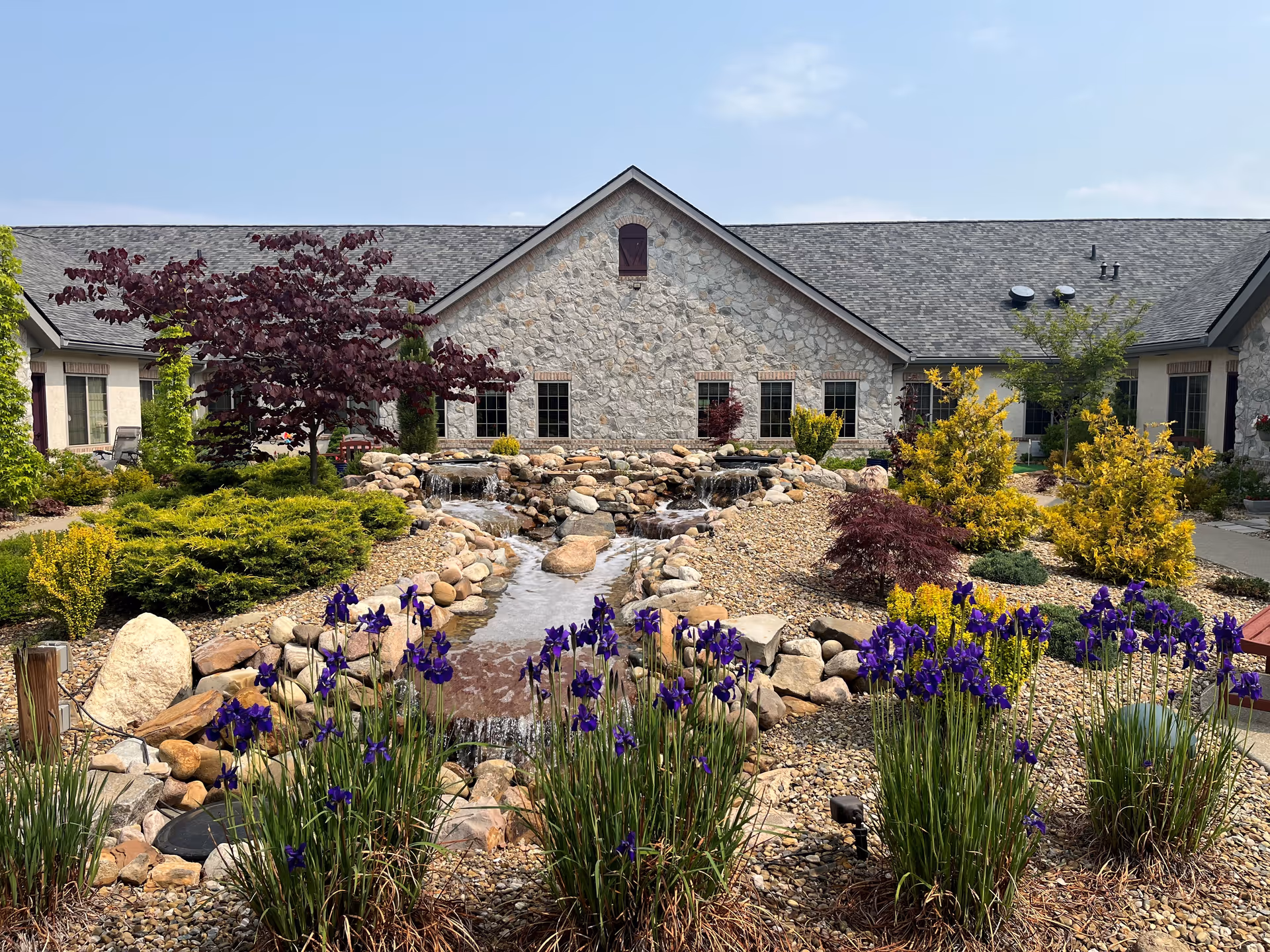 A landscaped outdoor garden area with a small rock waterfall and stream flowing through the center. Purple flowers and various green and yellow shrubs surround the water feature. In the background is a stone building with multiple windows and a gray shingled roof under a clear blue sky.
