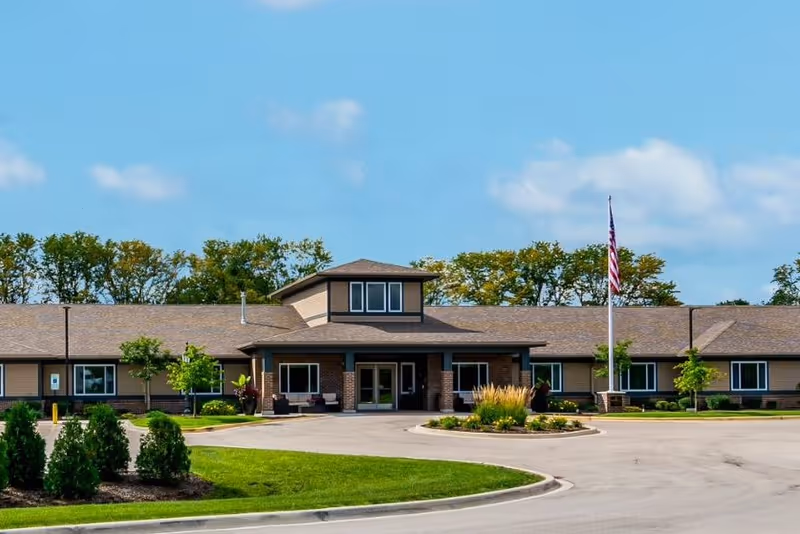 Front exterior view of Harvester Place facility with a single-story building, a covered entrance, an American flag on a flagpole, landscaped greenery, and a clear blue sky.