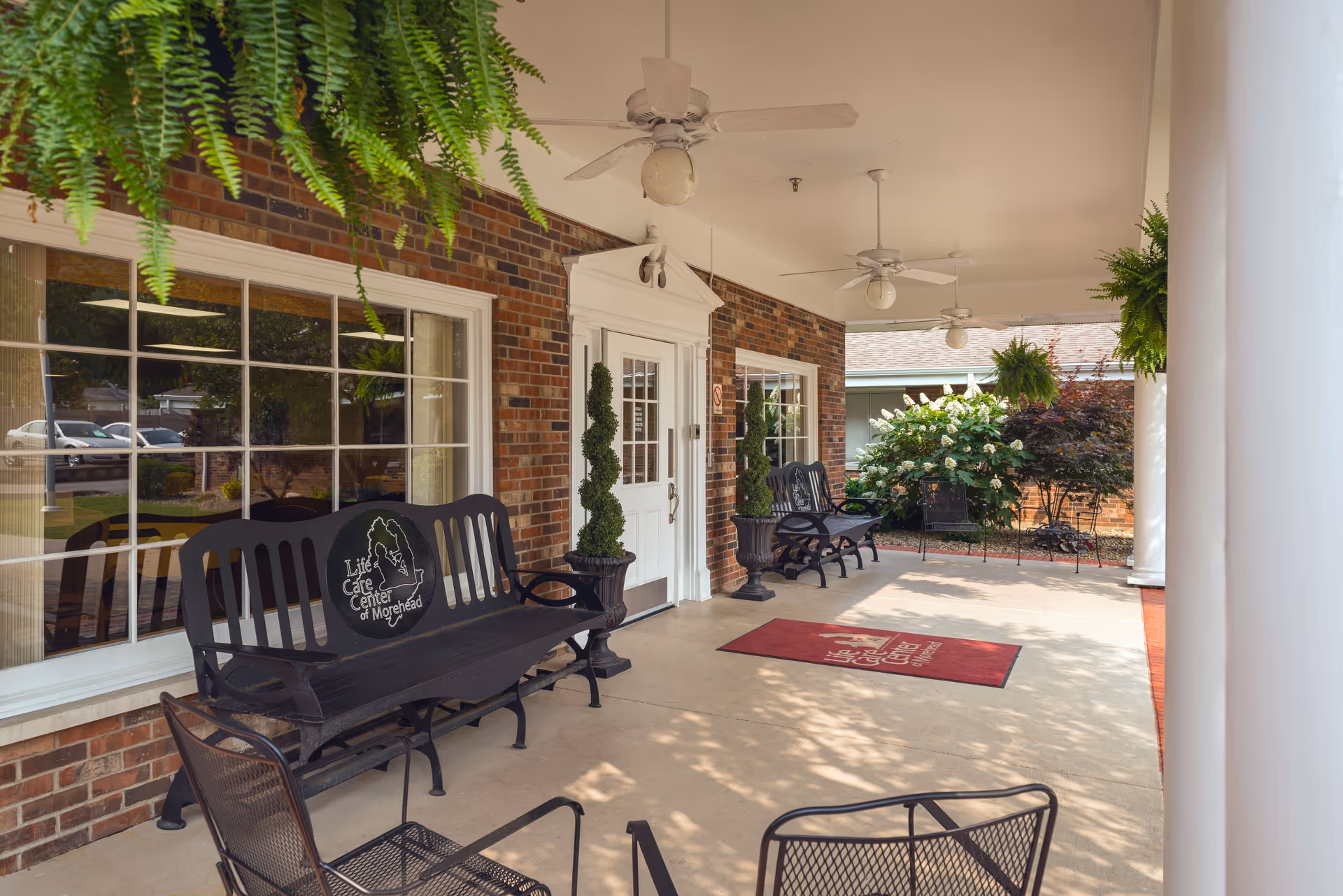 Covered outdoor patio area at Life Care Center of Morehead with black benches, hanging ferns, potted plants, ceiling fans, and a red welcome mat near the entrance door.