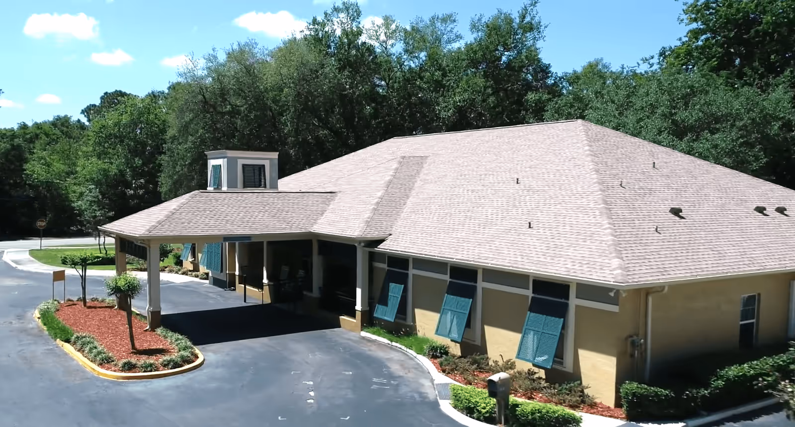 Front exterior of a single-story memory care building with a covered vehicle drop-off, teal shutters, and landscaped grounds.