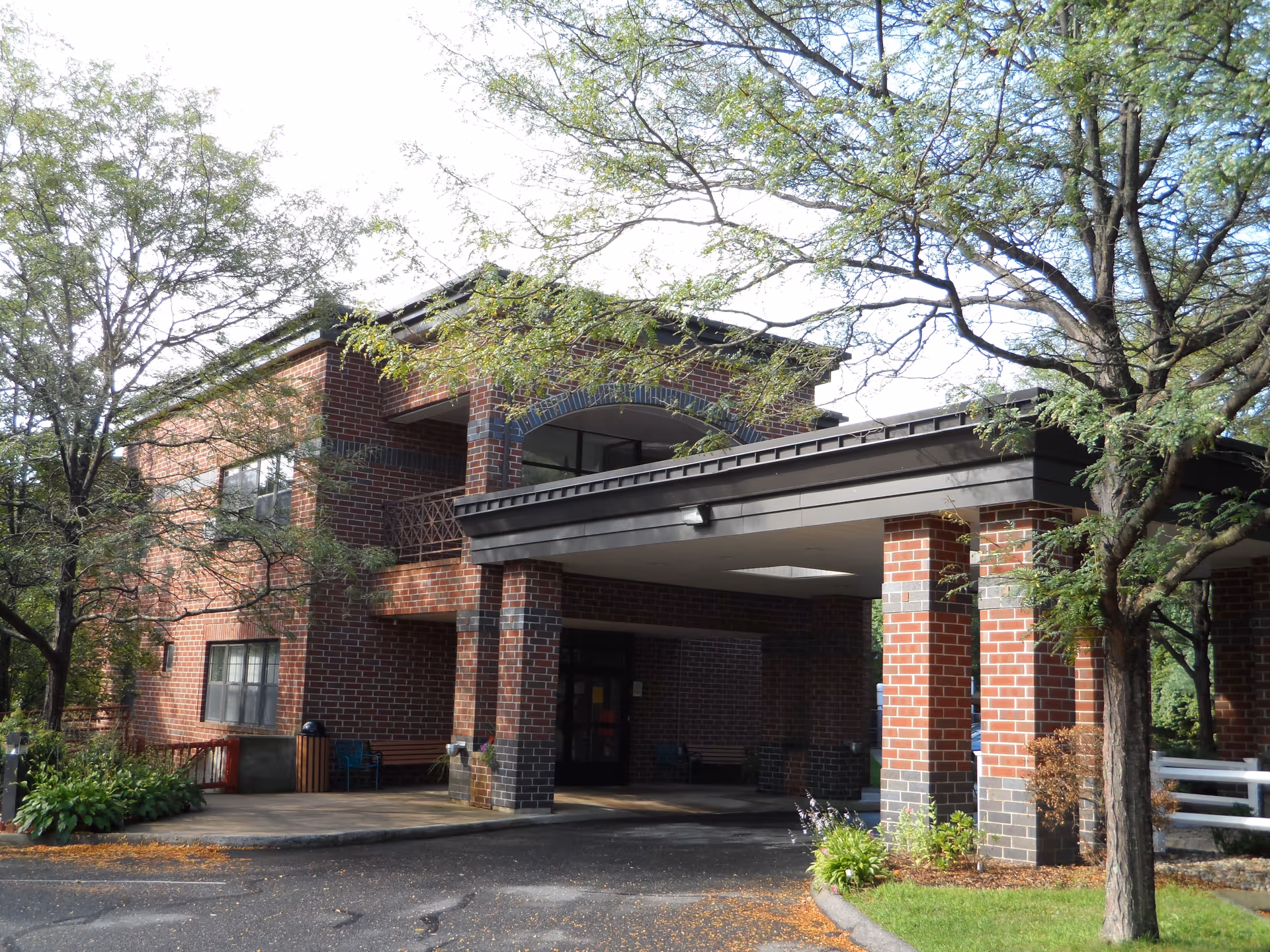 Exterior view of a two-story brick building with a covered entrance supported by brick columns. Trees with green leaves surround the building, and there is a paved driveway leading to the entrance. Some benches and plants are visible near the entrance area.