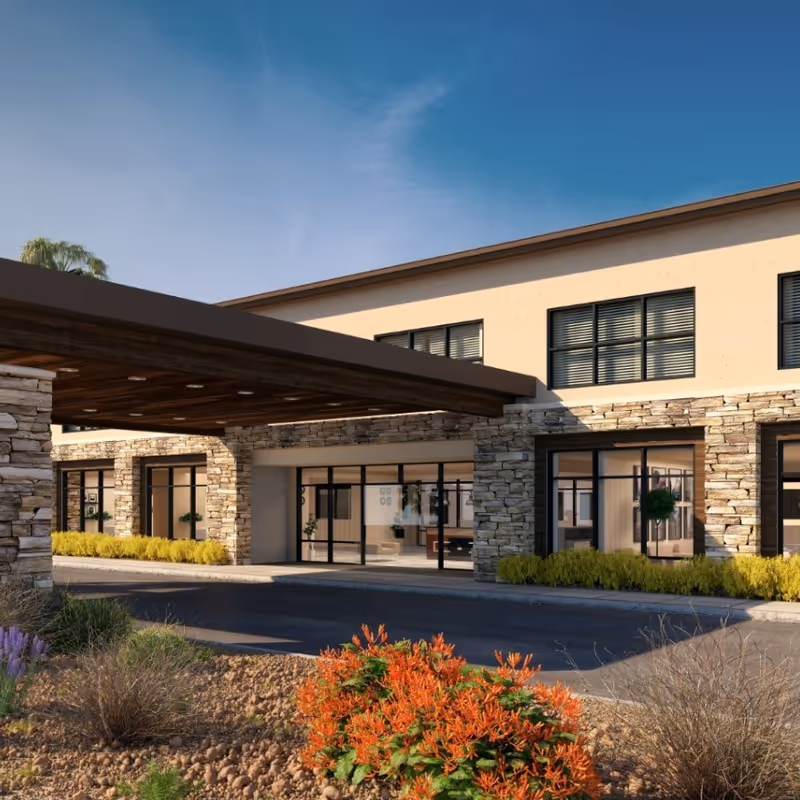 Exterior view of Carnegie Heights at Henderson senior living facility showing a covered entrance with stone pillars, large windows, and landscaped plants including orange flowers and shrubs under a clear blue sky.