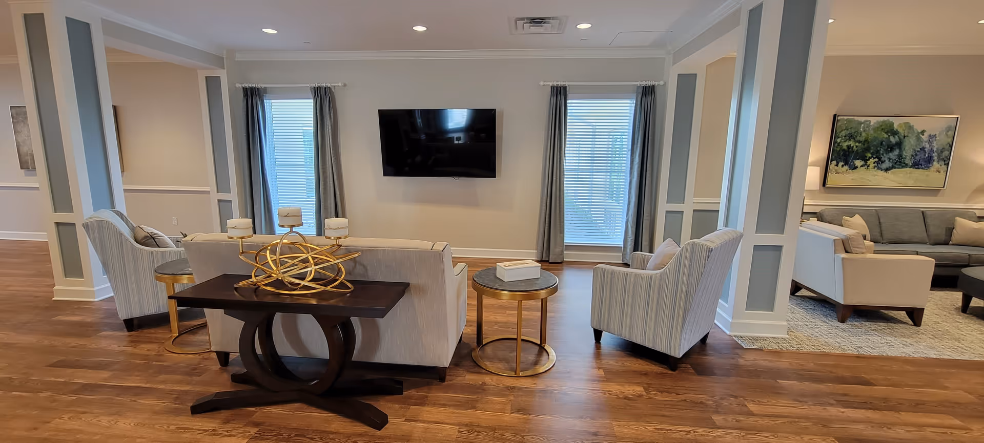A spacious and well-lit living room area with wooden flooring, featuring a beige sofa, two striped armchairs, a dark wooden console table with a decorative gold candle holder, and a round side table. A flat-screen TV is mounted on the wall between two windows with gray curtains. In the background, another seating area with a gray sofa, beige armchair, and a large framed painting is visible.